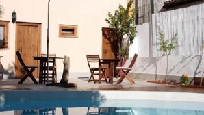 Un espace piscine agrémenté de chaises longues et d’une table, entouré d’un bâtiment doté d’un toit en tuiles et d’un volet vert.