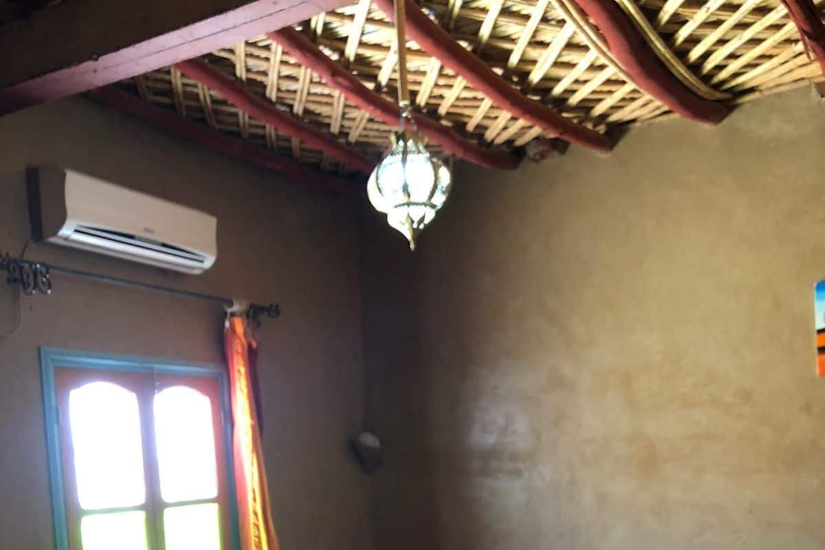 Ein Schlafzimmer mit Bett, kleinem Tisch, Fenster mit Vorhängen, Klimaanlage und einer Pendelleuchte.