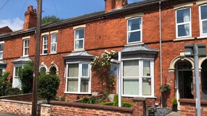 A red brick terraced house with white-framed windows, a bay window with white curtains, and a small garden with flowers and plants. - 202 Guesthouse (Lincoln)