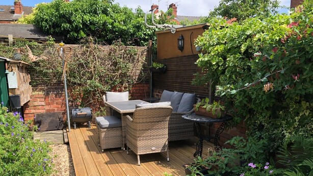A wooden deck with outdoor furniture, surrounded by greenery and a brick wall. - 202 Guesthouse (Lincoln)