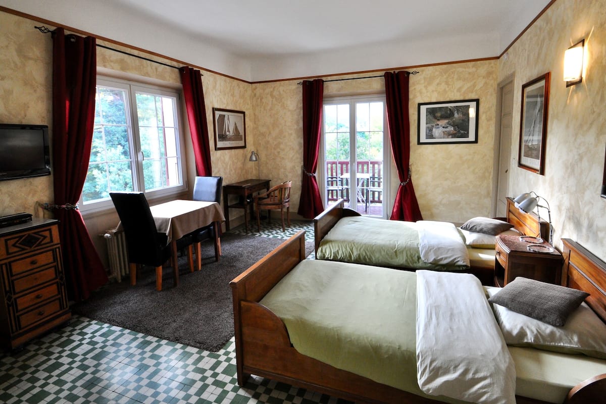 A hotel room with two beds, a desk, a chair, a TV, and a balcony with red curtains.