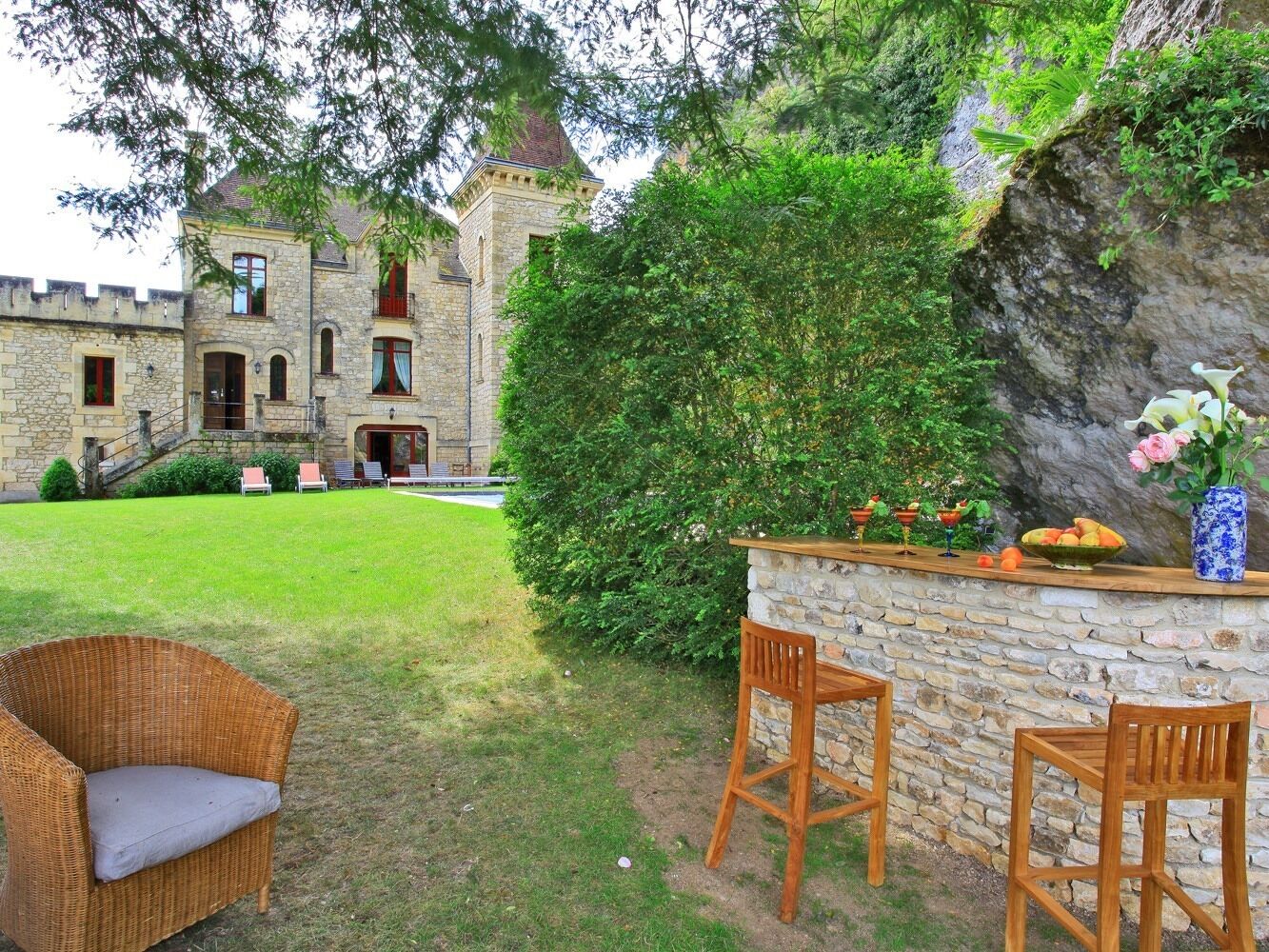 A stone building with a tower, a grassy yard, and a stone bar area with chairs and a vase of flowers.