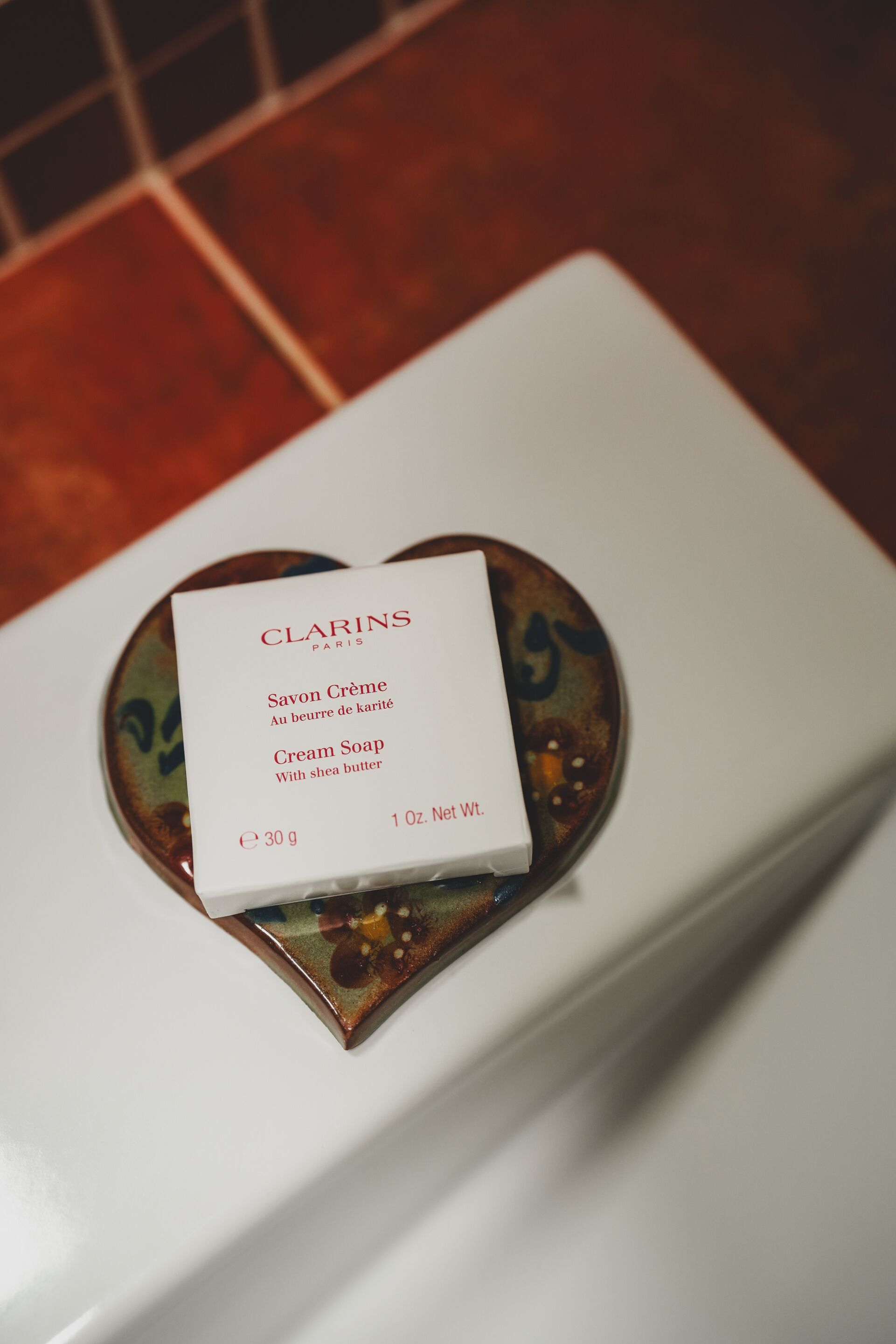 Una caja blanca con el rótulo 'CLARINS PARIS', que contiene el texto 'Savoir Crème' y 'Cream Soap', sobre una superficie blanca con un plato decorativo debajo.