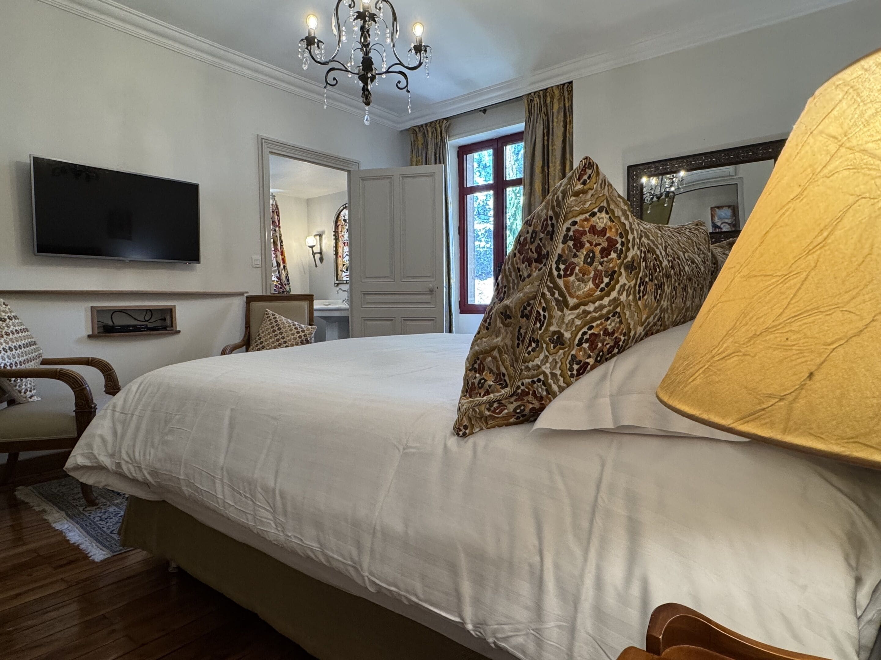 A hotel room with a large bed, a television, a chandelier, a mirror, and a window with curtains.