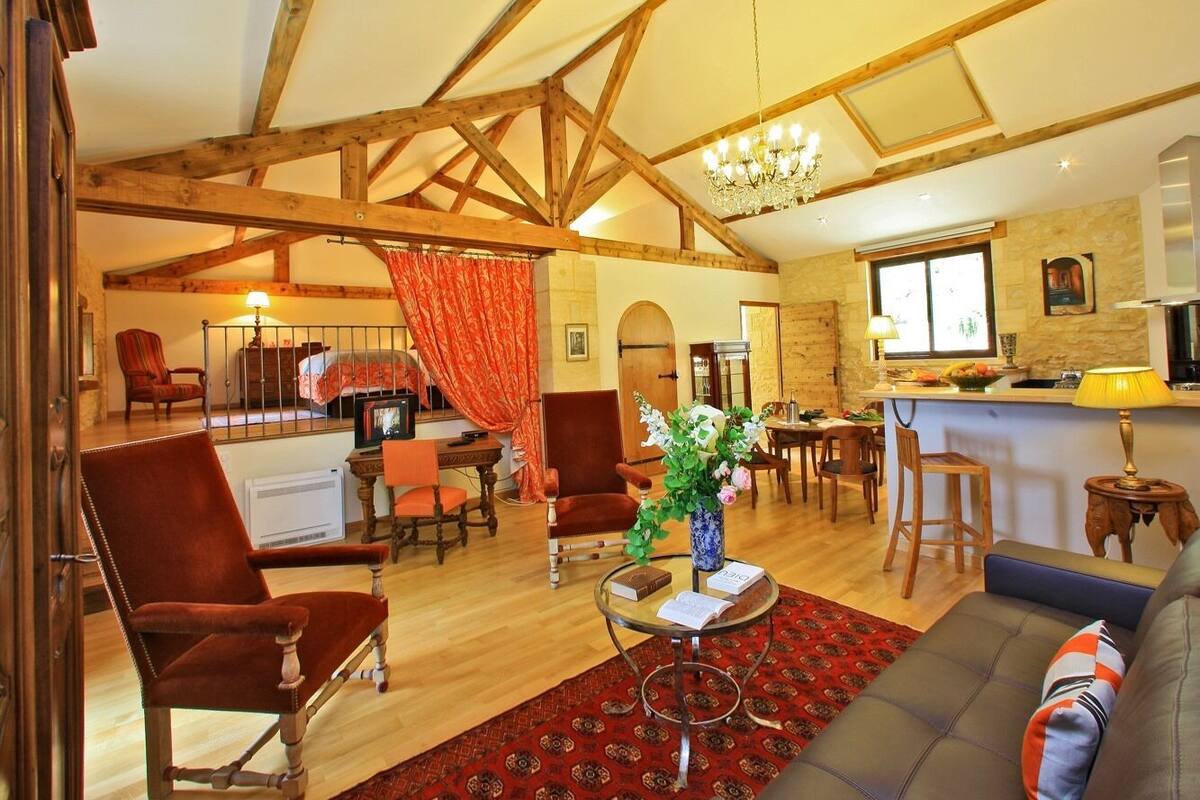 A spacious living area with a high ceiling, wooden beams, a chandelier, a dining area with a table and chairs, and a kitchen visible through an open door.