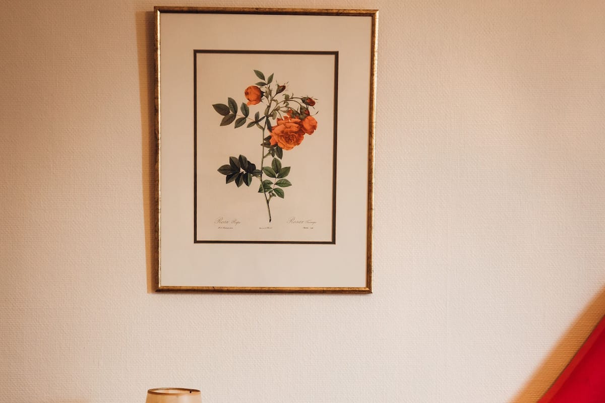 A neatly arranged bedroom with a bed, a bedside table with a lamp, a framed botanical print on the wall, and a colorful pillow.