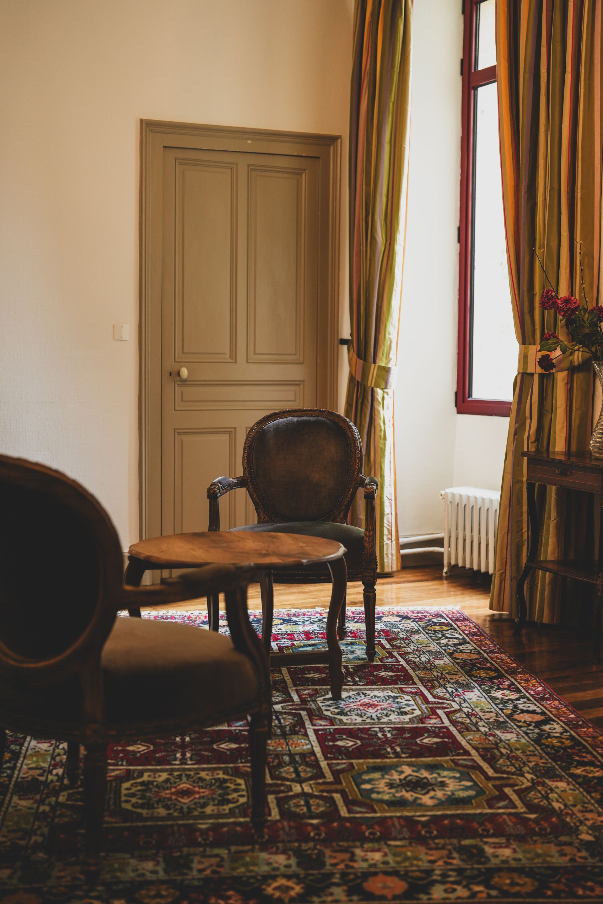 A room with a wooden chair, a table, a patterned rug, a window with curtains, and a radiator.