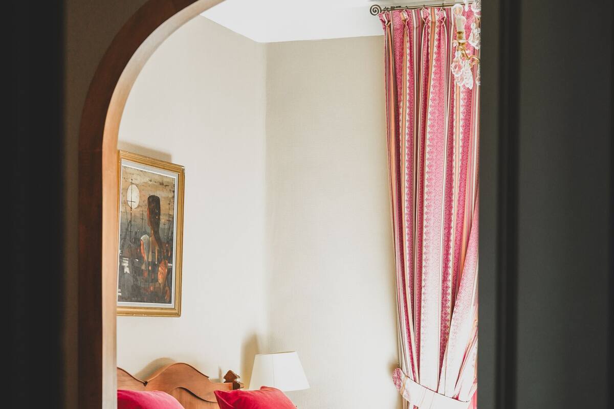 A bedroom with a bed, pillows, a nightstand, a lamp, a chair, a table, and a vase with flowers.