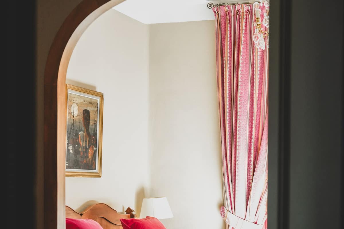 A bedroom with a bed, pillows, a nightstand, a lamp, a chair, a table, and a vase with flowers.