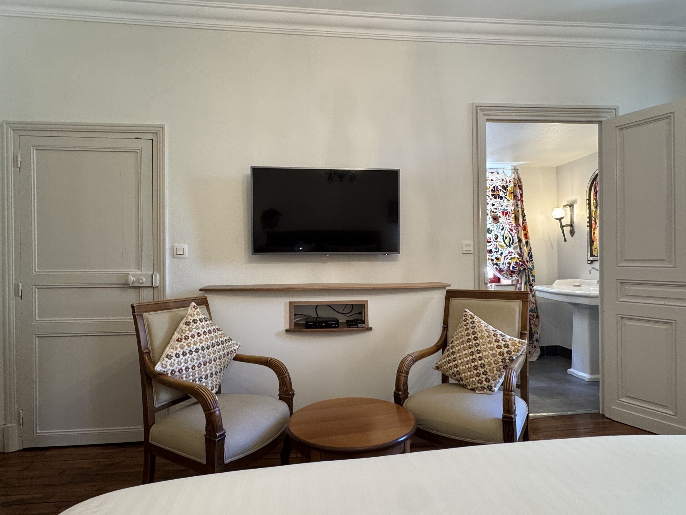 A hotel room with a TV mounted on the wall, two wooden chairs with patterned cushions, a small wooden table, and a bathroom visible through an open door.