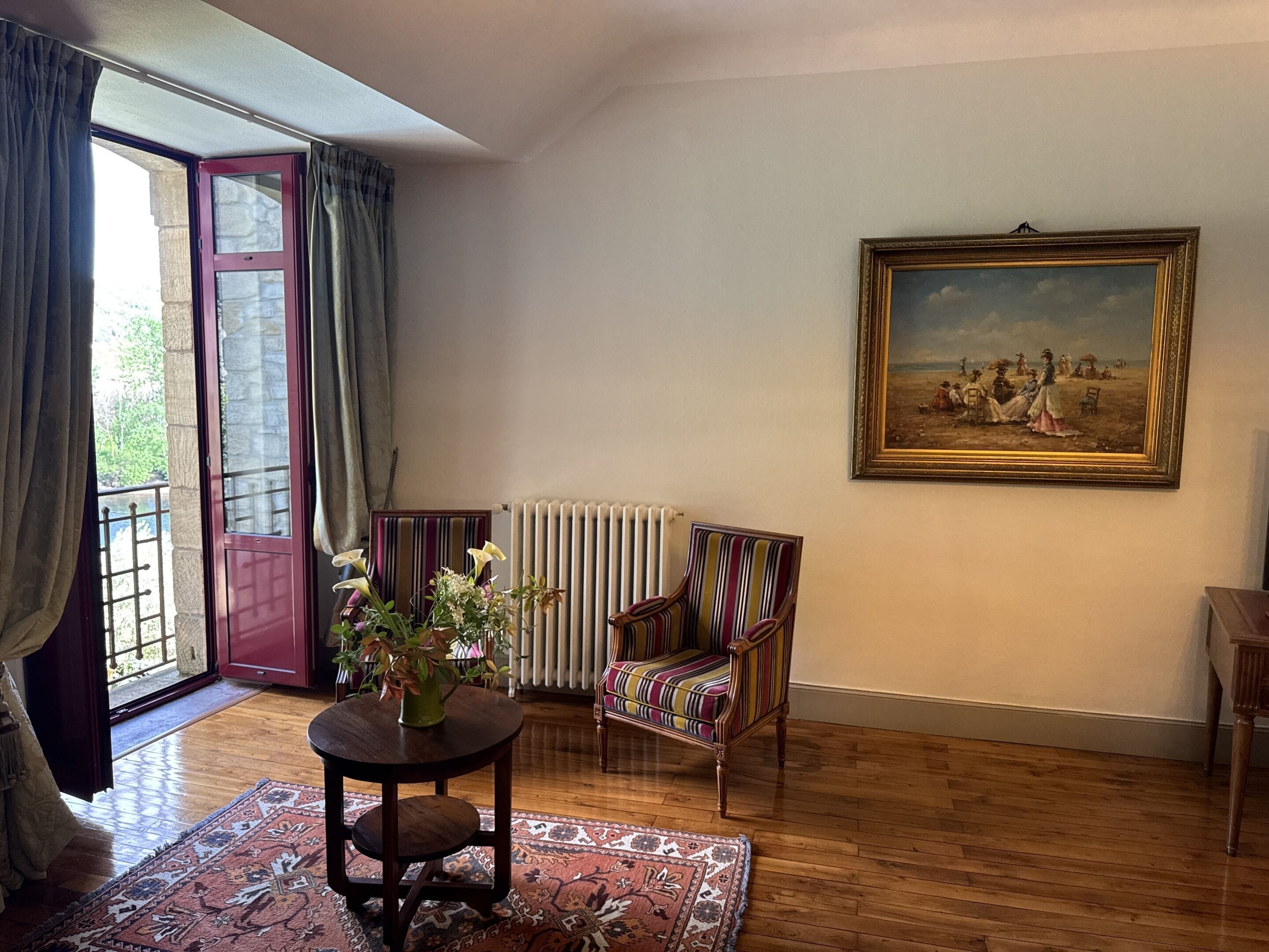 A room with a wooden floor, a radiator, a striped armchair, a table with a vase of flowers, a painting on the wall, and a door with curtains.