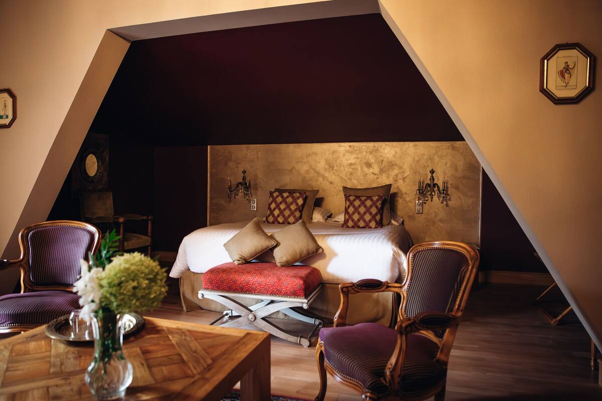 A triangular room with a bed, two chairs, a small table, and a vase with flowers.