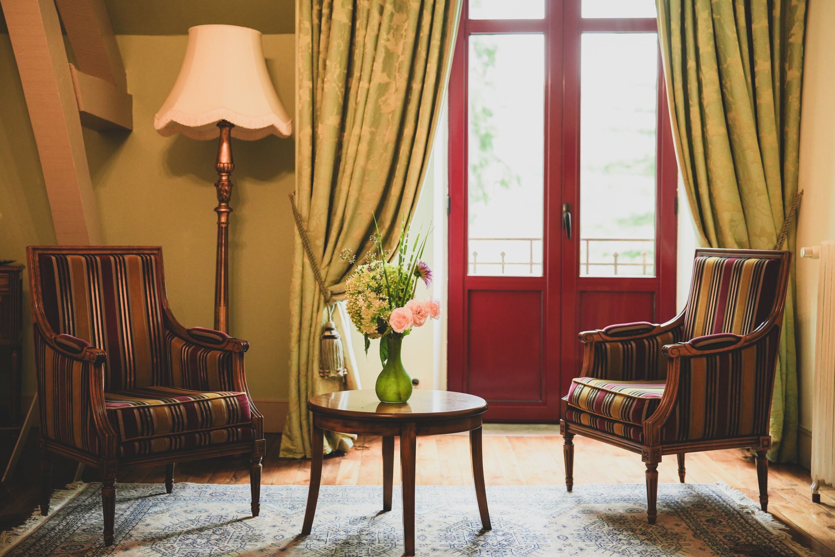 A room with two striped armchairs, a round wooden table with a vase of flowers, a lamp, and a red door with curtains.