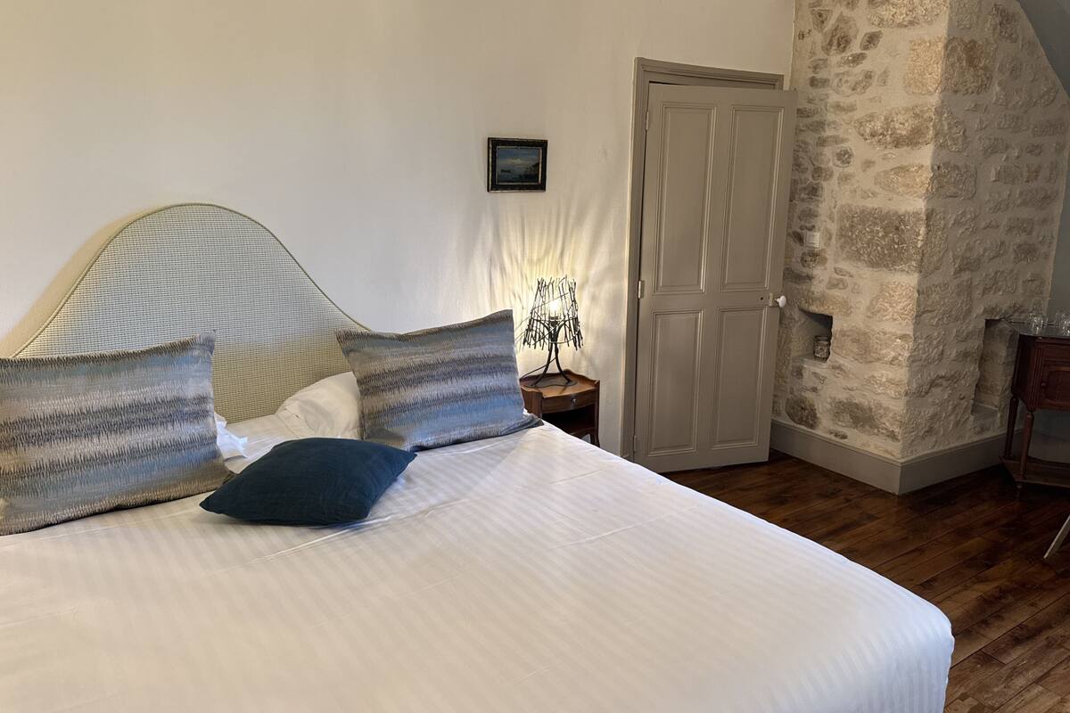 A bedroom with a bed, pillows, a nightstand, a wall-mounted lamp, a door, and a stone wall.