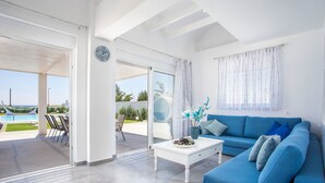 A modern living room with a blue sofa, a white coffee table, and a view of a pool and outdoor area. - White Passion Villa (Rhodes)