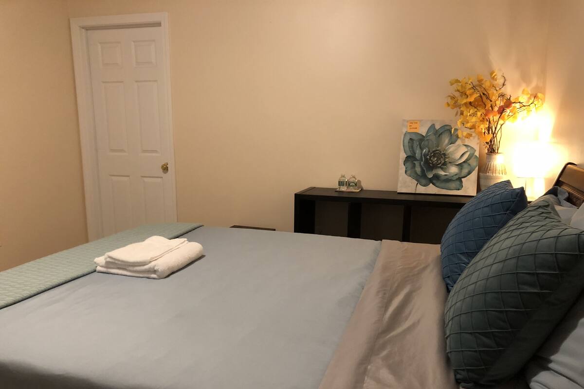 A neatly made bed with a folded white towel, a bedside table with a lamp, and a wall-mounted artwork.