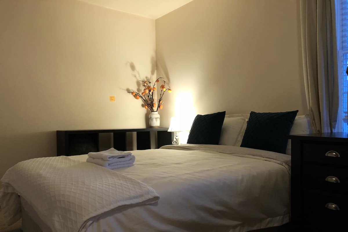 A hotel room with a neatly made bed, a bedside table with a lamp, and a vase with dried flowers on the wall.