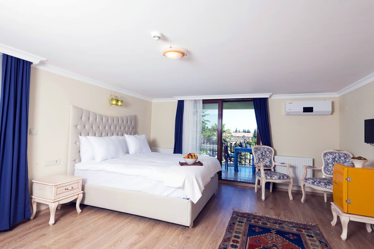 A hotel room with a large bed, a small bedside table, a chair, a yellow cabinet, and a balcony with a table and chairs.