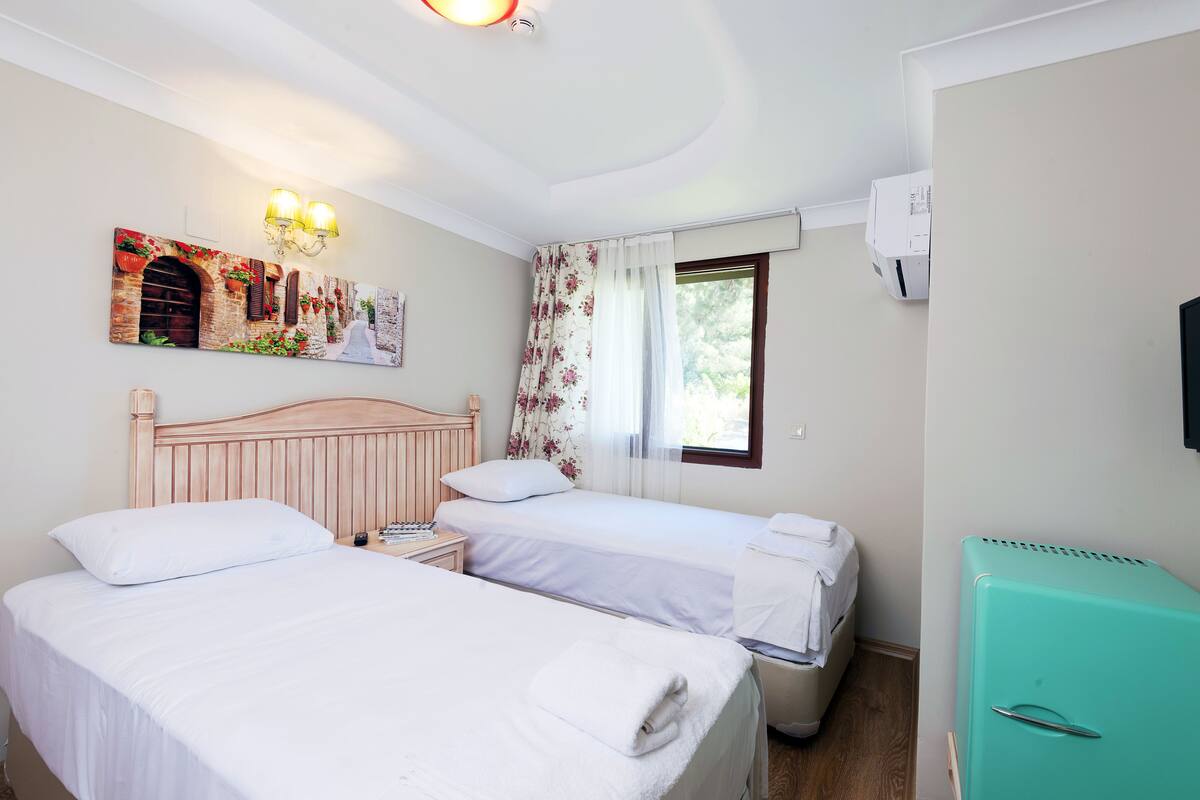 A hotel room with two beds, a wooden headboard, a wall-mounted artwork, a window with floral curtains, and a teal-colored refrigerator.