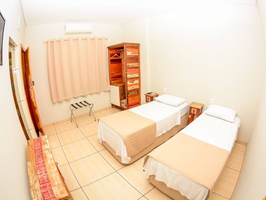 Habitación de hotel con dos camas, un armario de madera, una mesita, una silla y una ventana con cortinas.