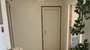 A spacious hallway with a wicker chair and a cushioned bench, a potted plant, and a door with a sign indicating check-in times. - Cunda Paradise Otel (Ayvalik)