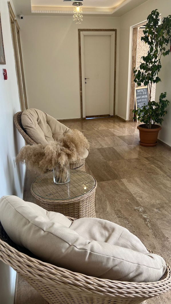 Un pasillo amplio con una silla de mimbre y un banco acolchado, una planta en maceta y una puerta con un cartel que indica los horarios de registro.