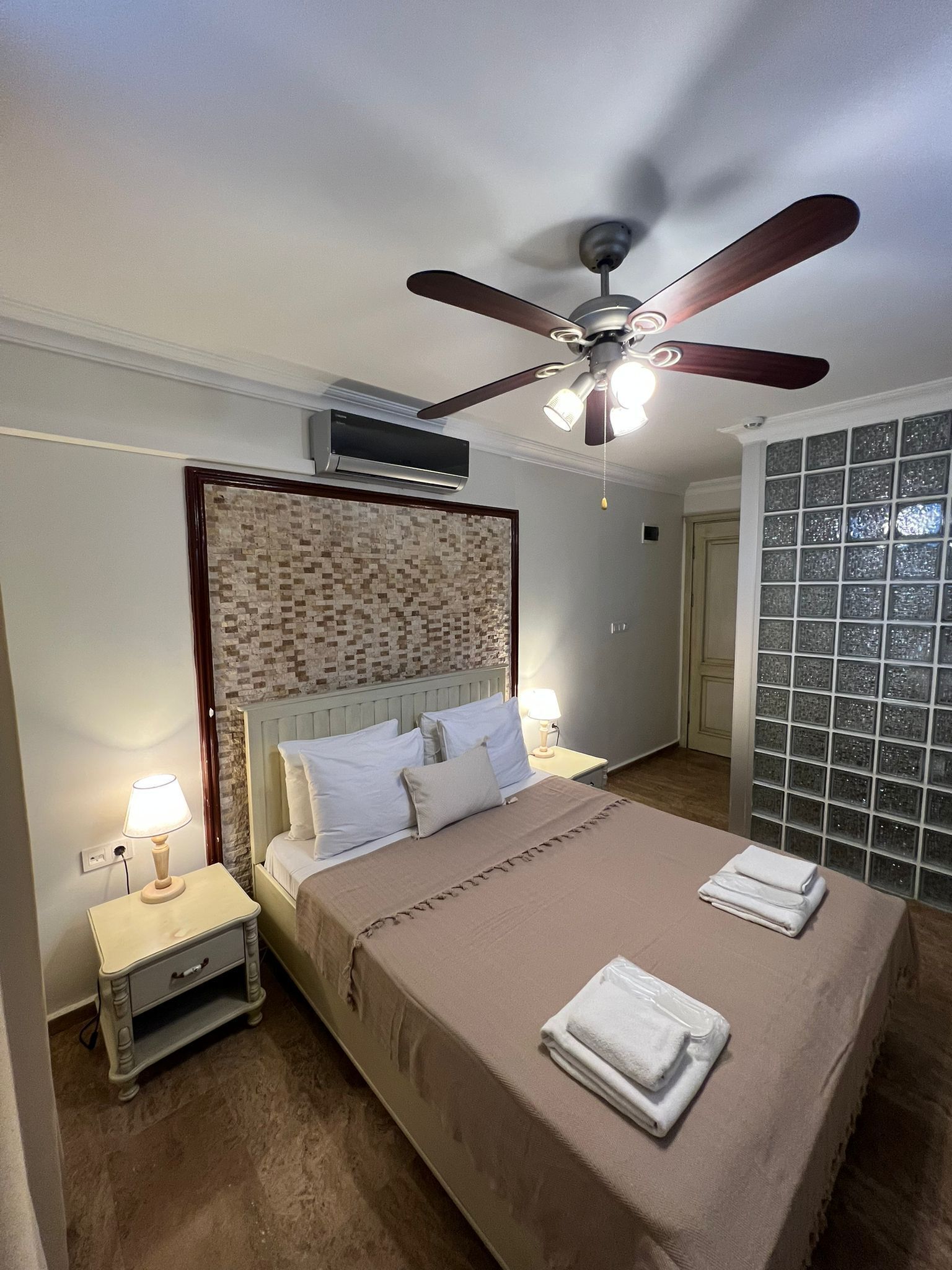Un dormitorio con cama, ventilador de techo, mesita de noche, lámpara y una pared con diseño de mosaicos.