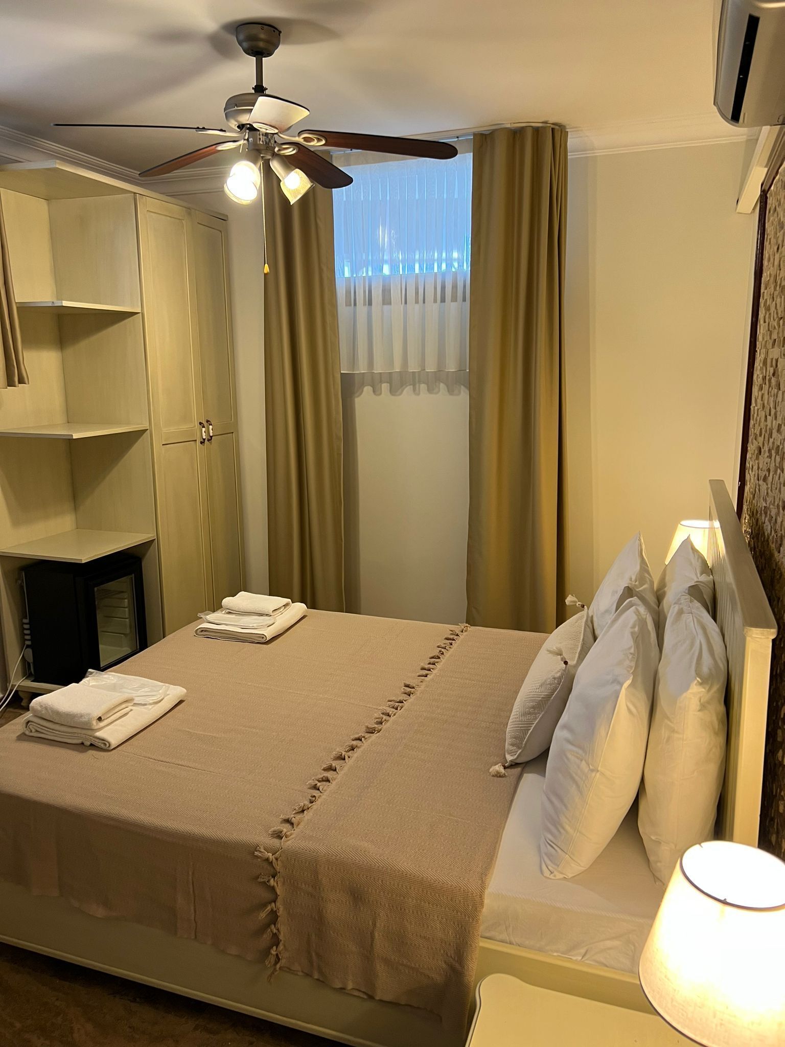 A hotel room with a bed, a ceiling fan, a window with curtains, and a bedside lamp.
