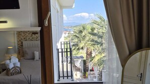 A balcony with a view of palm trees and a distant landscape, a bed with a white headboard, and a small table with a lamp and decorative items. - Cunda Paradise Otel (Ayvalik)