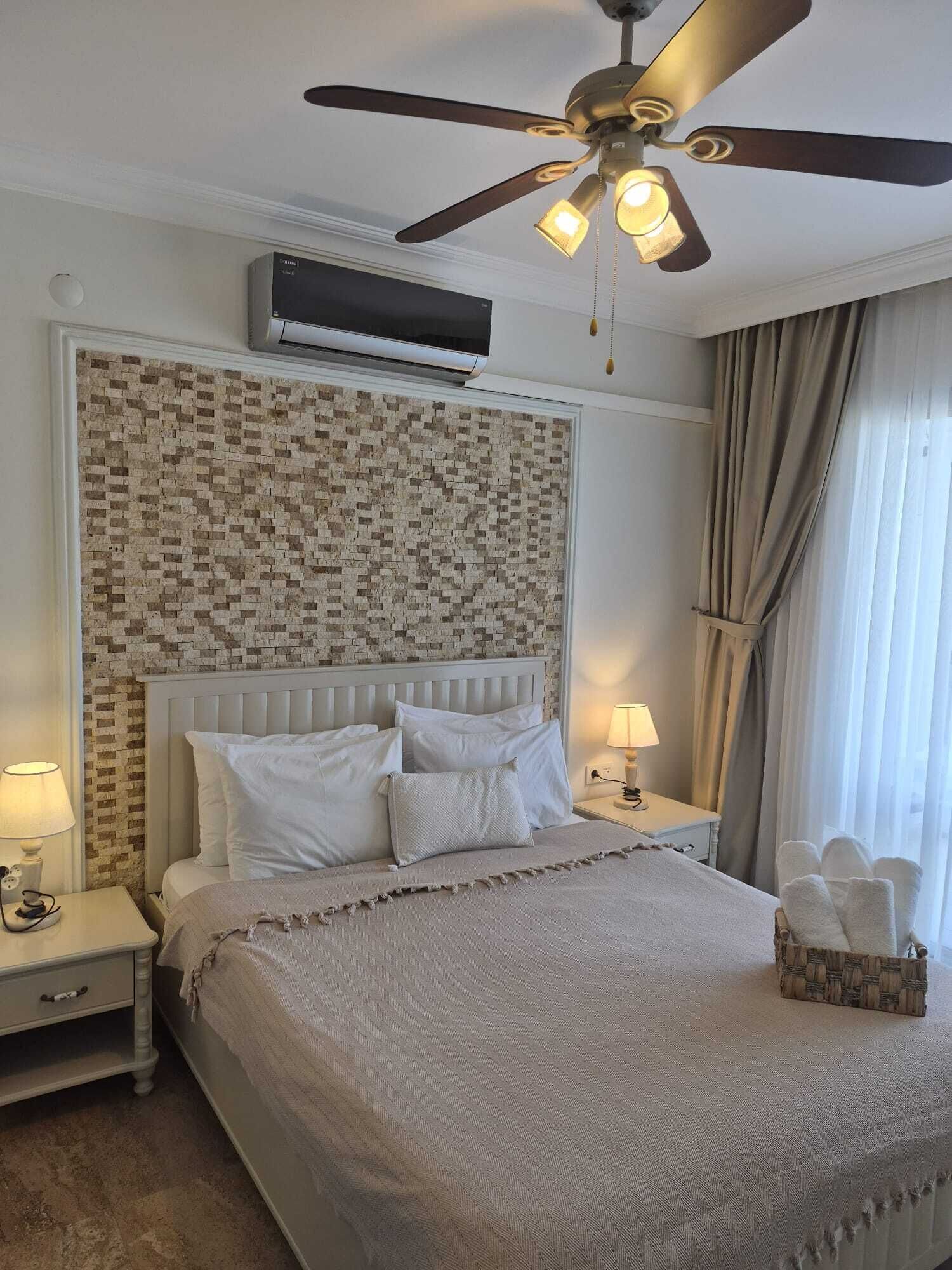 A hotel room with a bed, a ceiling fan, an air conditioning unit, a bedside lamp, and a window with curtains.