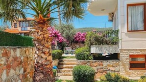 A well-maintained garden with a palm tree, stone walls, and a pathway leading to a building with a balcony. - Cunda Paradise Otel (Ayvalik)