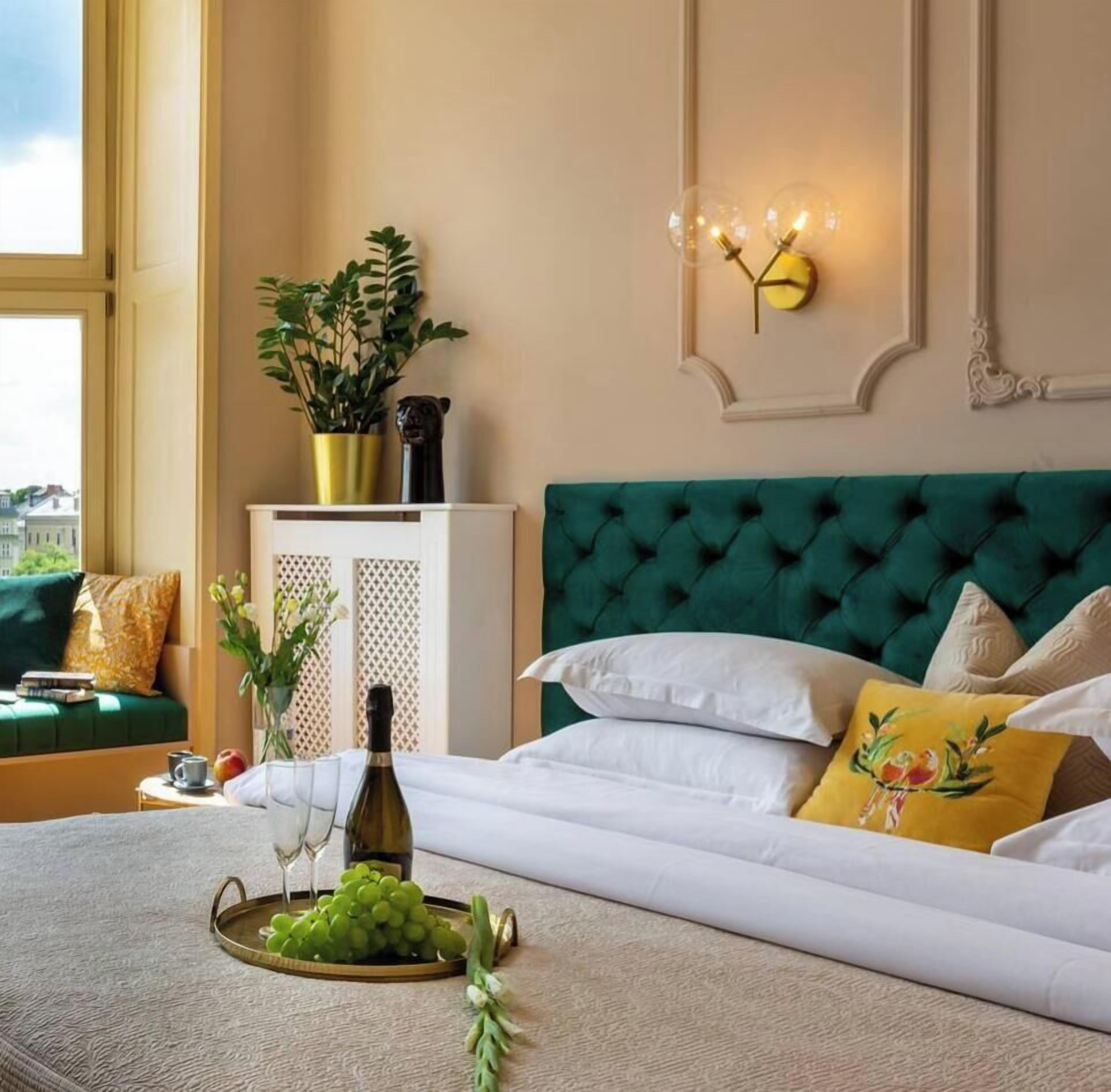 Une chambre d’hôtel avec une tête de lit recouverte d’un tissu vert, une table de chevet avec une bouteille et des verres, un plateau de raisins et une vue sur la ville par la fenêtre.