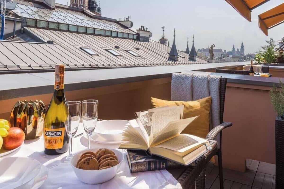 Eine Dachterrasse mit einem Tisch, der für eine Mahlzeit gedeckt ist, darunter Früchte, eine Weinflasche und ein Buch.