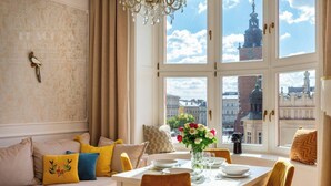 Ein Essbereich mit Tisch für zwei Personen, einer Couch mit Kissen, einer Kronleuchter und Blick auf Stadtgebäude durch große Fenster.
