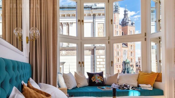 Ein Zimmer mit einem großen Erkerfenster, einem türkisfarbenen Sofa und Blick auf historische Gebäude.