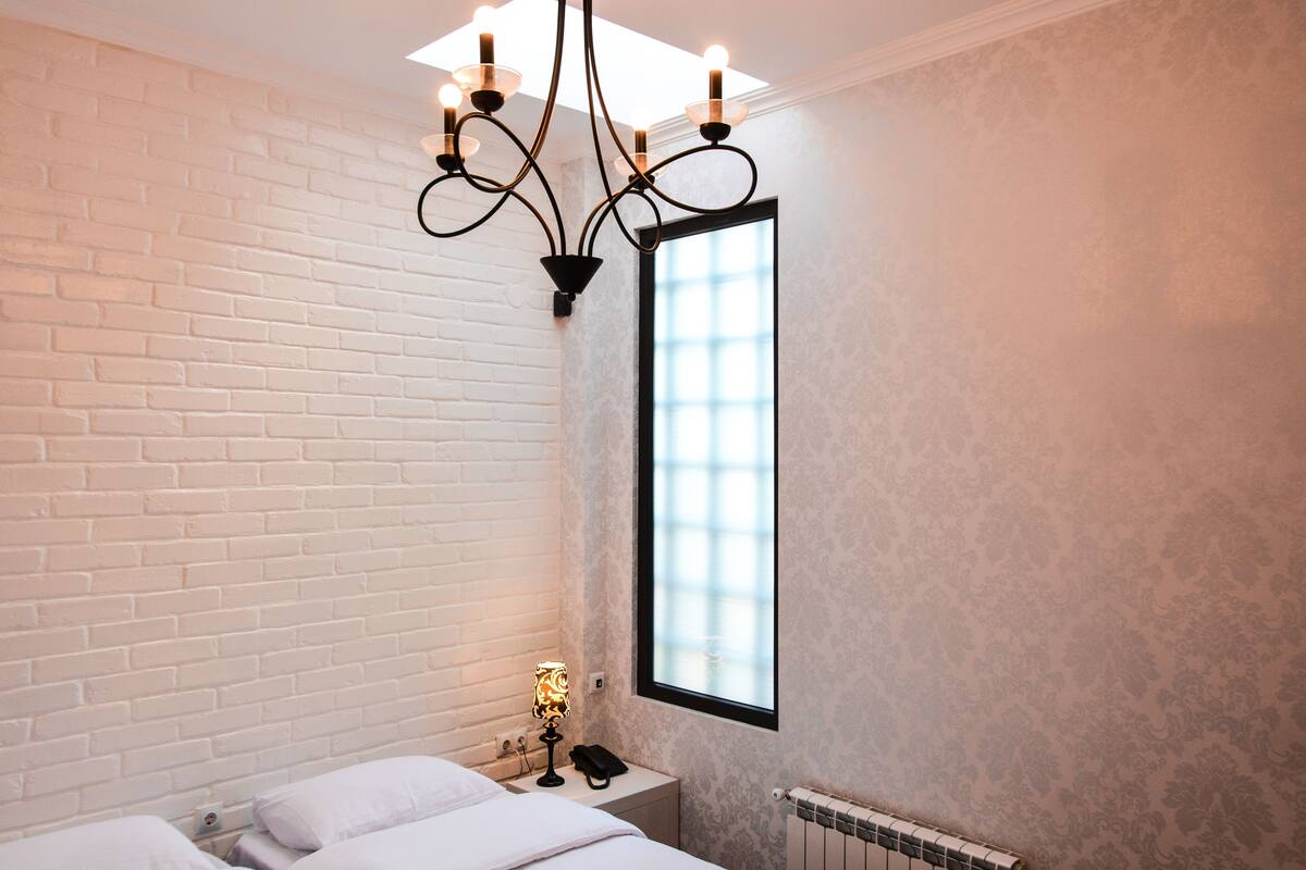 Een hotelkamer met een witte baksteenmuur, een groot raam, een bed met wit beddengoed, een nachtkastje met een lamp en een radiator.