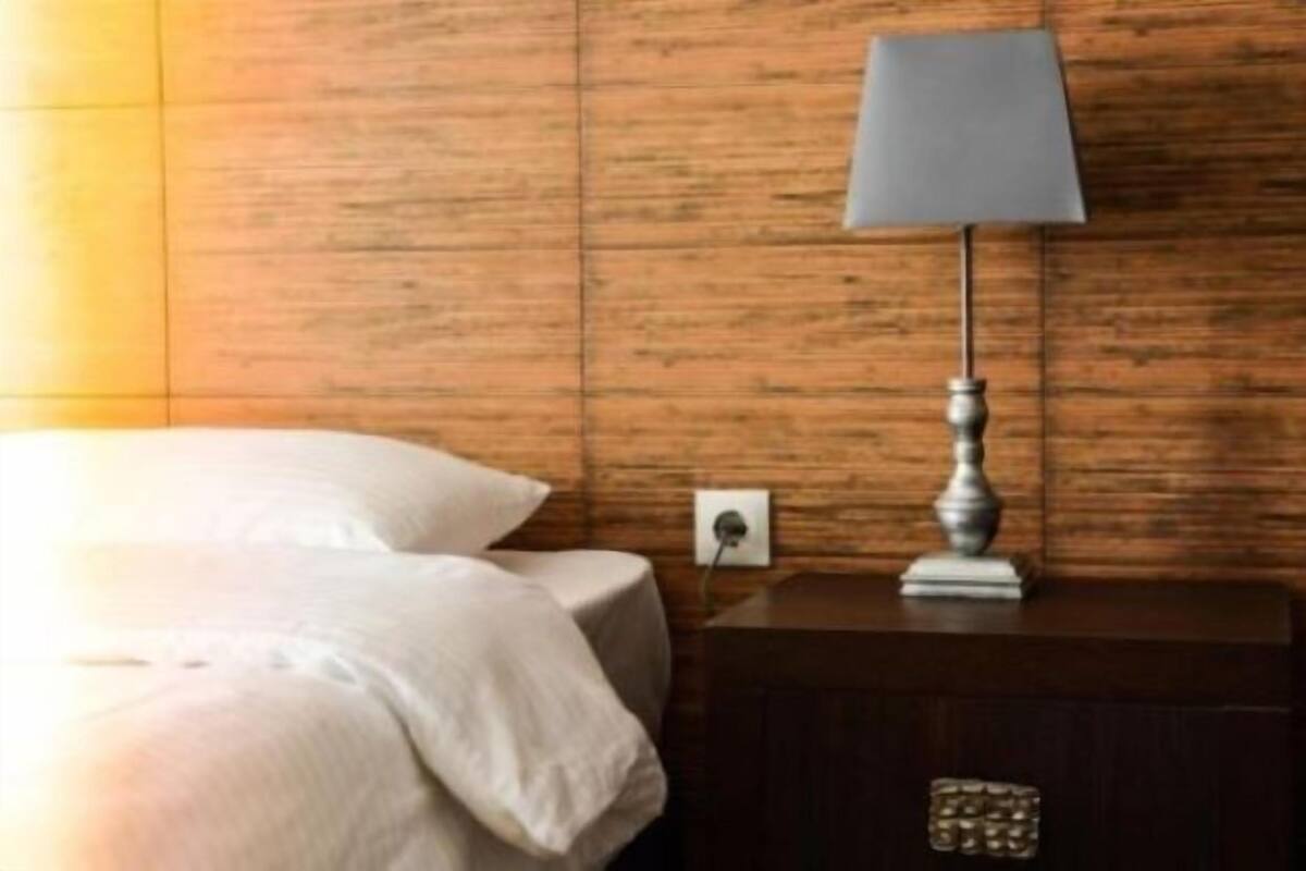 Een net opgemaakt bed met een witte deken en kussens, een houten nachtkastje met een lamp en een stopcontact.