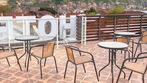 Una terraza con mesas y sillas, con vistas a un paisaje urbano que incluye colinas y una iglesia.