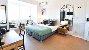 A modern bedroom with a large bed, a desk, and a chair. There is a window, a mirror, and an air conditioning unit. - Archisuites (Coimbra)