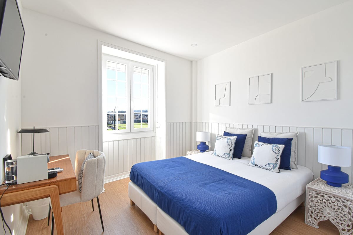 A bedroom with a bed, a desk, and a chair. There is a window with a view of buildings outside.