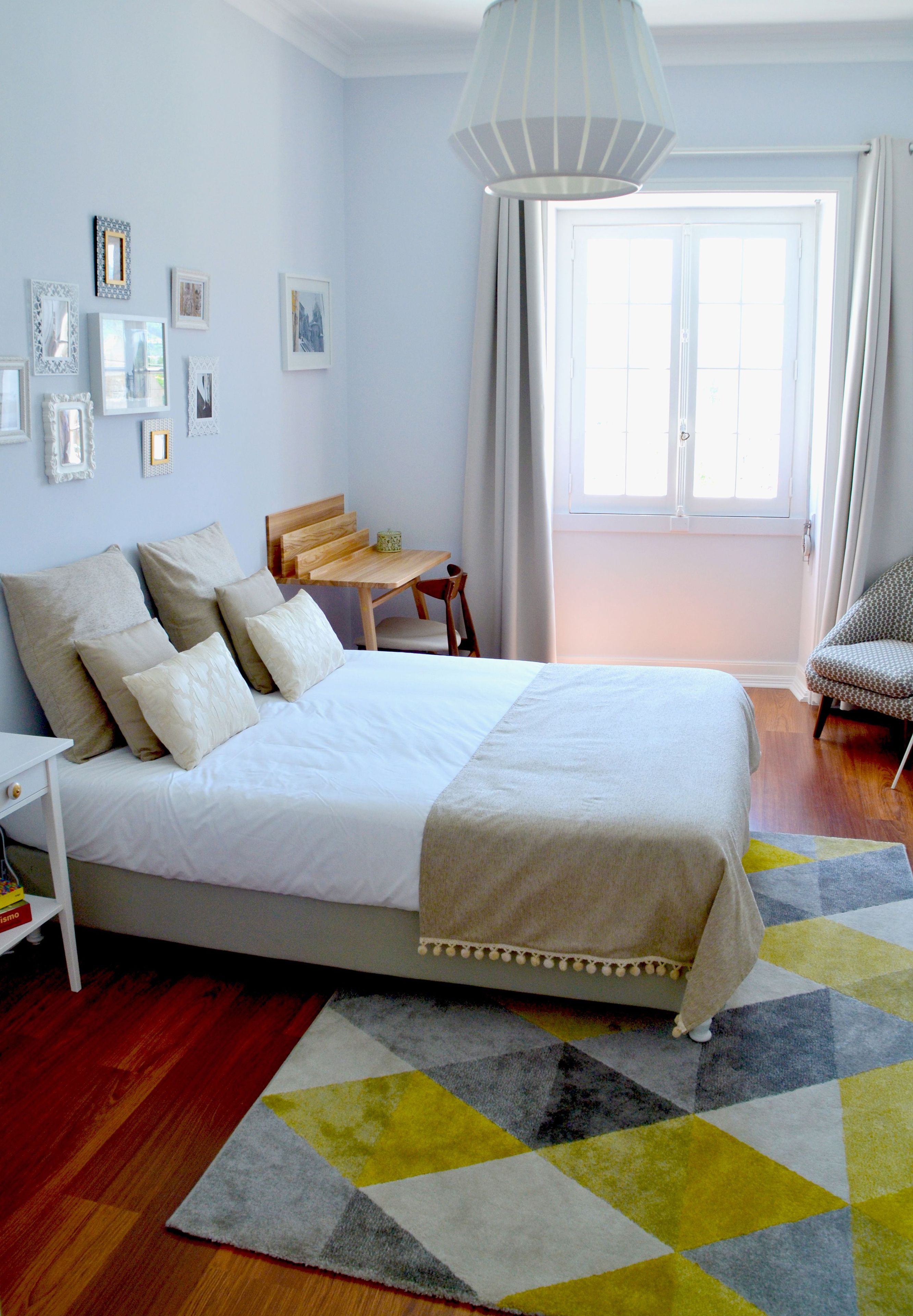 A bedroom with a bed, a wooden chair, a small table, and a large window with curtains.