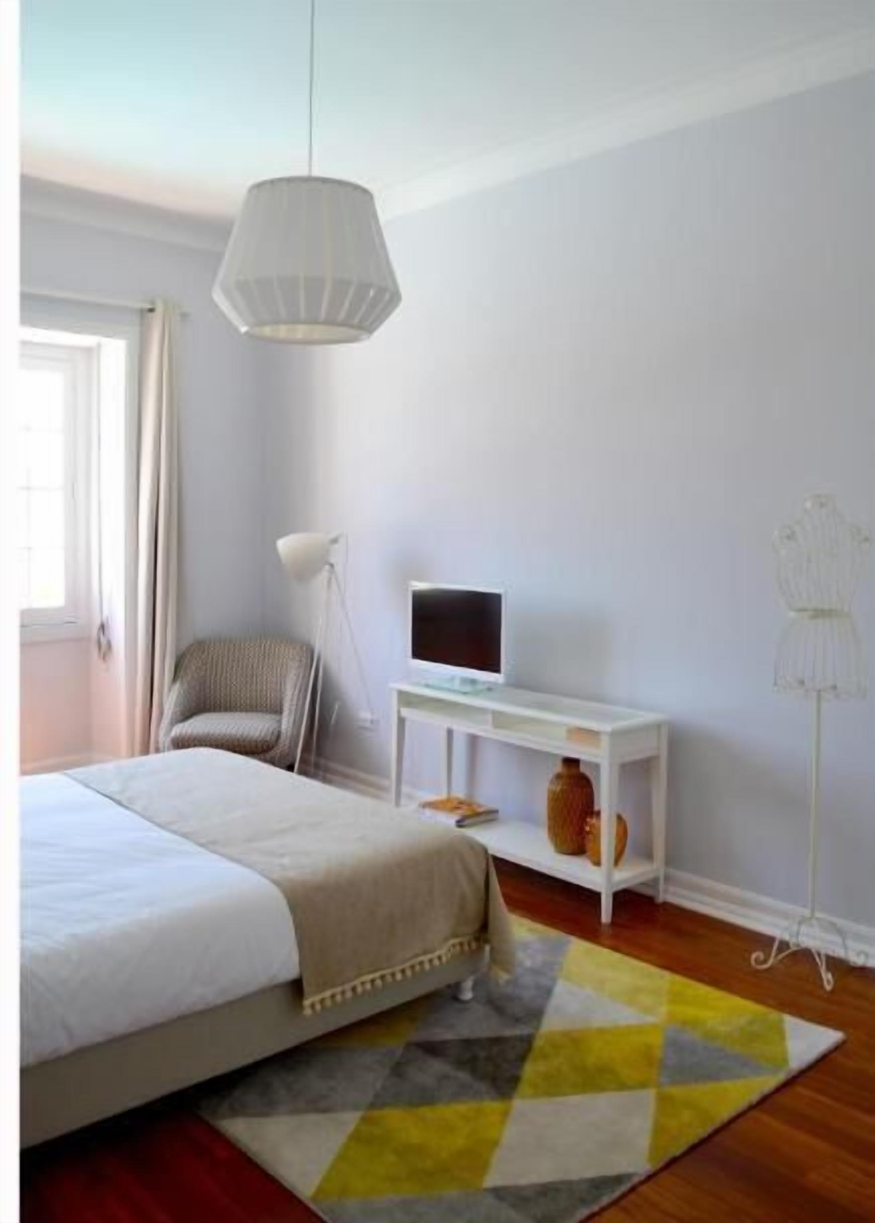 A bedroom with a bed, a TV on a white stand, a chair, and a floor lamp.