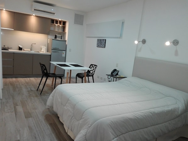 A compact hotel room with a bed, a small kitchenette, a dining table with chairs, and a phone on a side table.