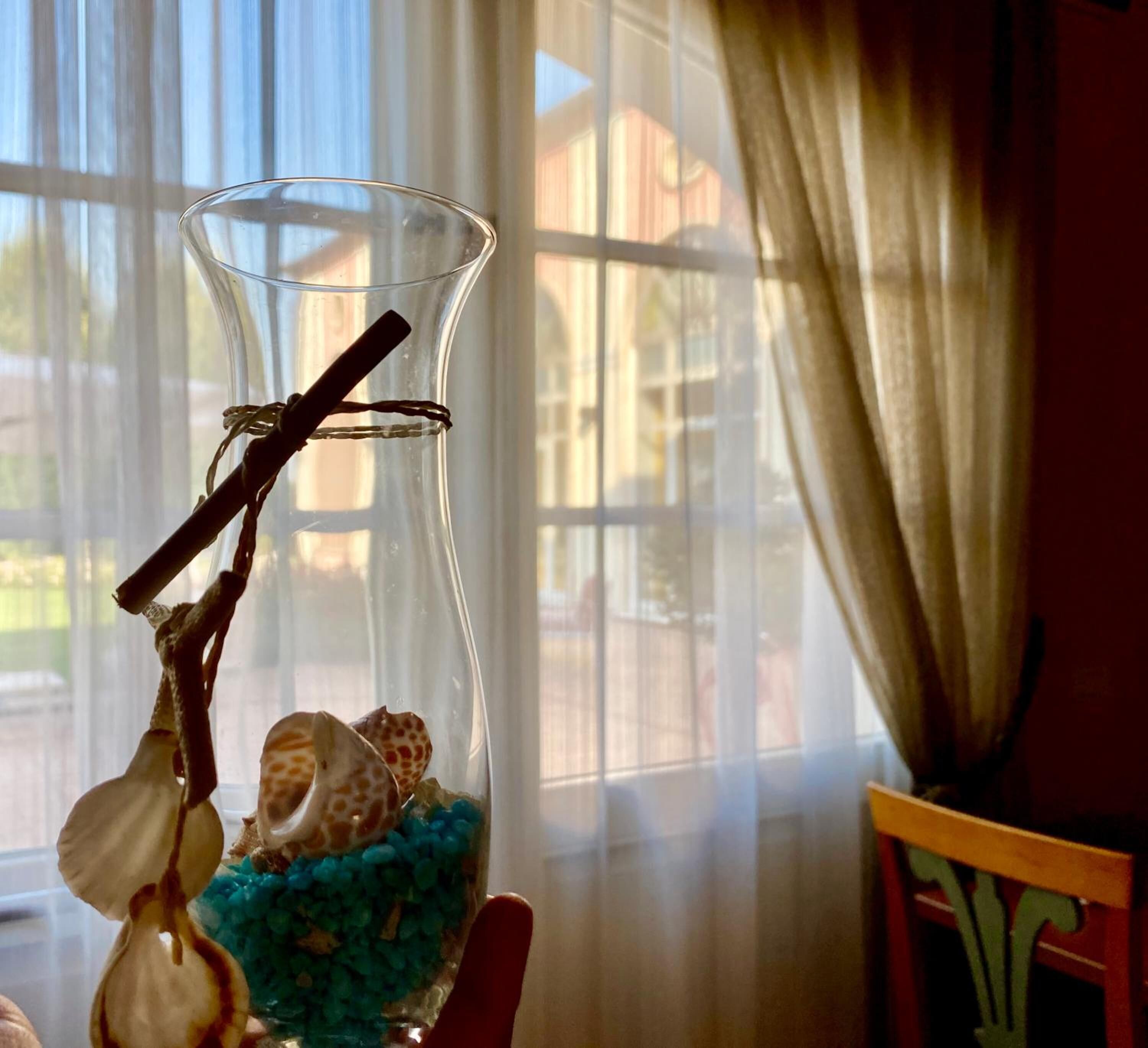 A clear glass vase with seashells and blue pebbles, a cinnamon stick, and a dried twig, placed on a table by a window with sheer curtains.