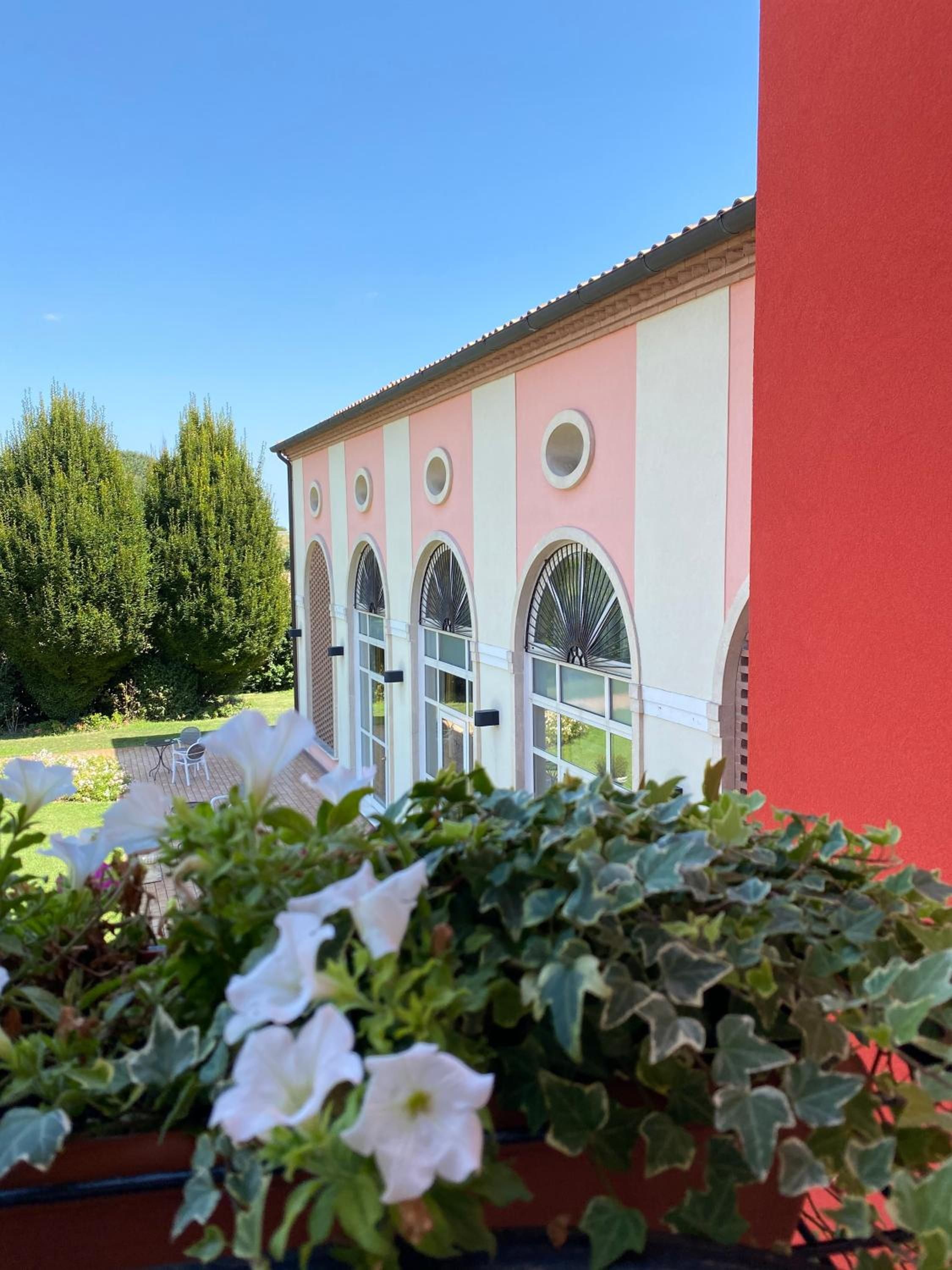 Un edificio con fachada rosa, ventanas arqueadas y un jardín con flores blancas y plantas verdes.
