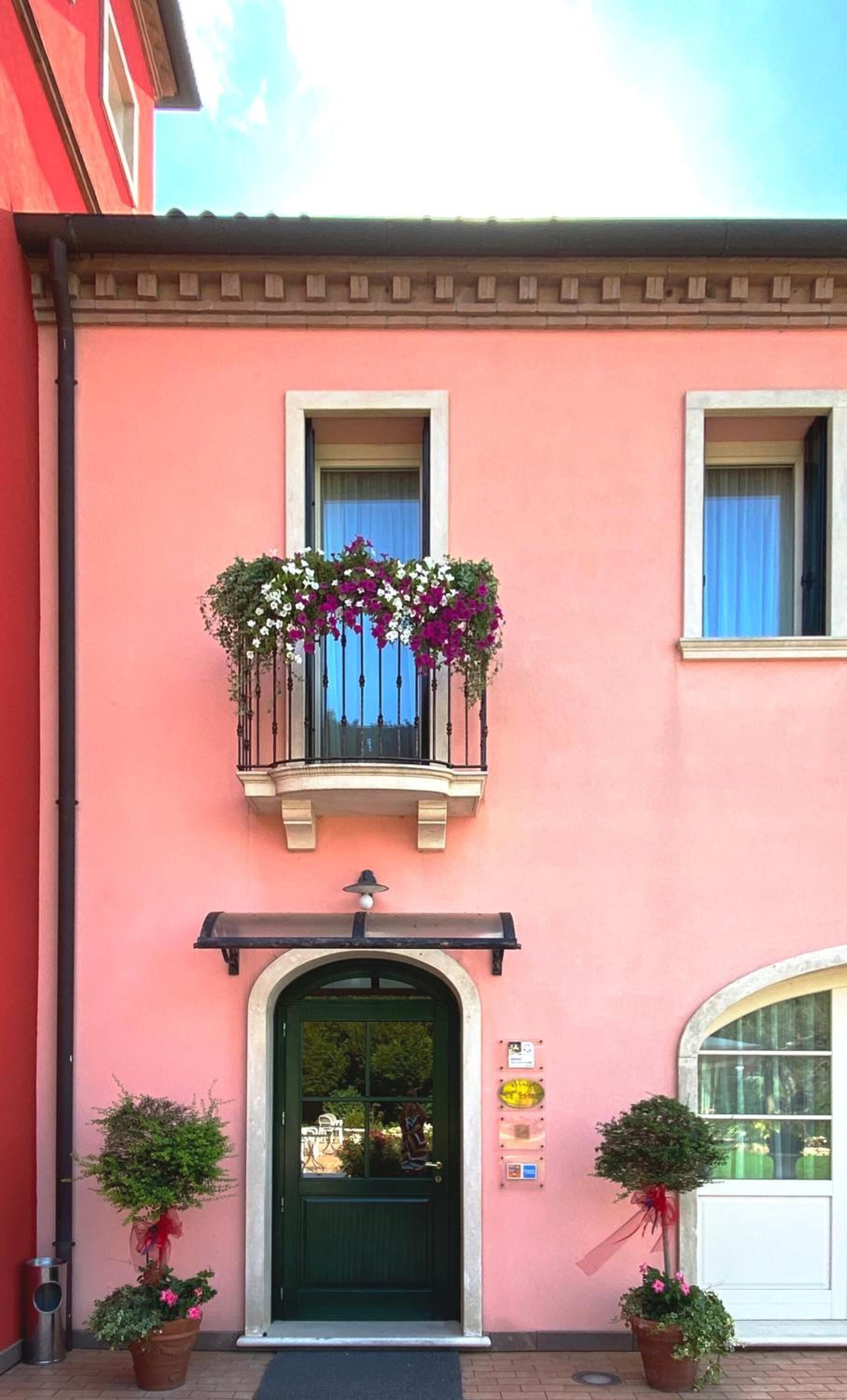 A pink building with a green door, a balcony with flowers, and two potted plants.