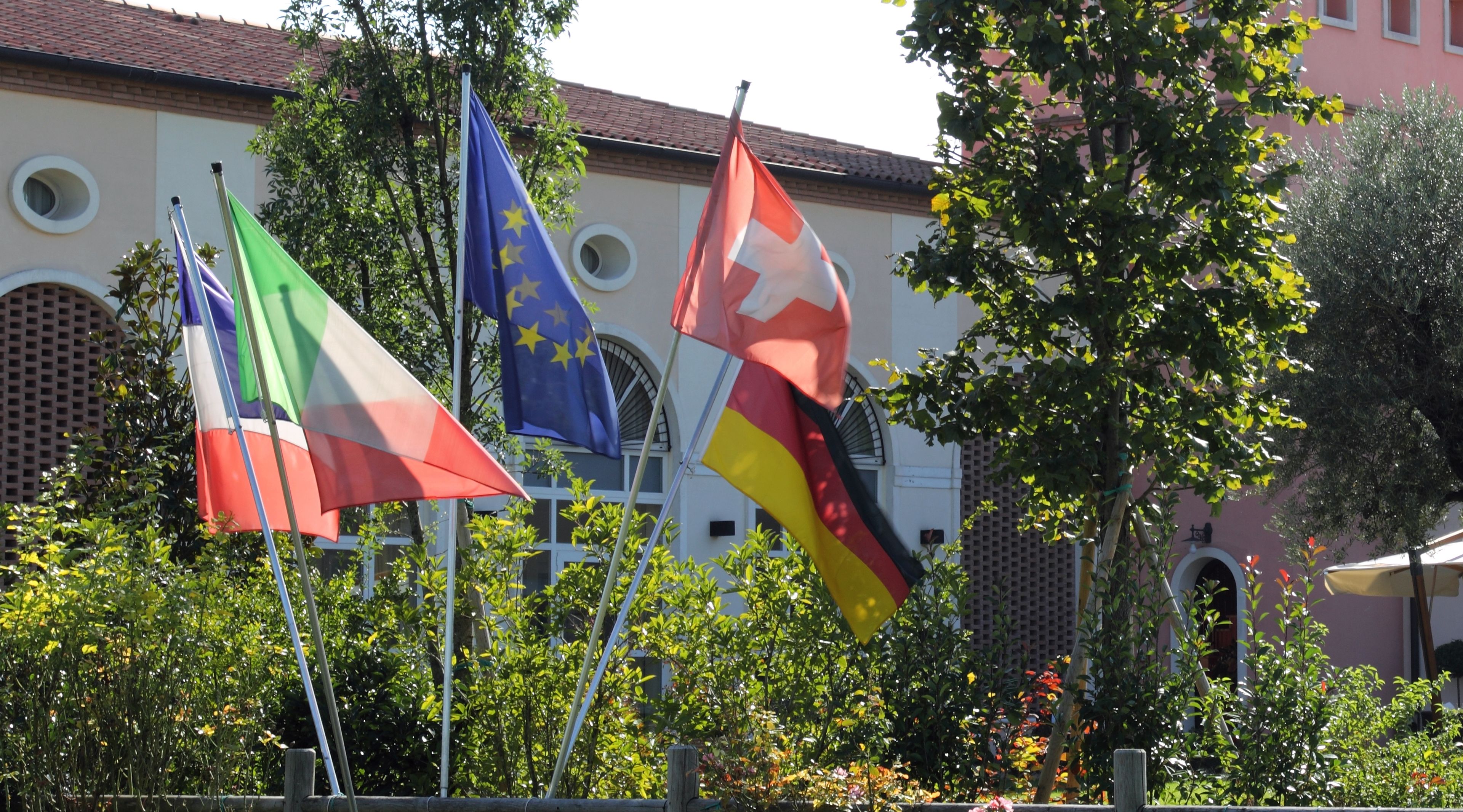 Banderas de Italia, Alemania y la Unión Europea frente a un edificio con ventanas arqueadas y techo de tejas.