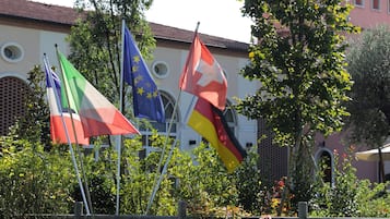 Banderas de Italia, Alemania y la Unión Europea frente a un edificio con ventanas arqueadas y techo de tejas.