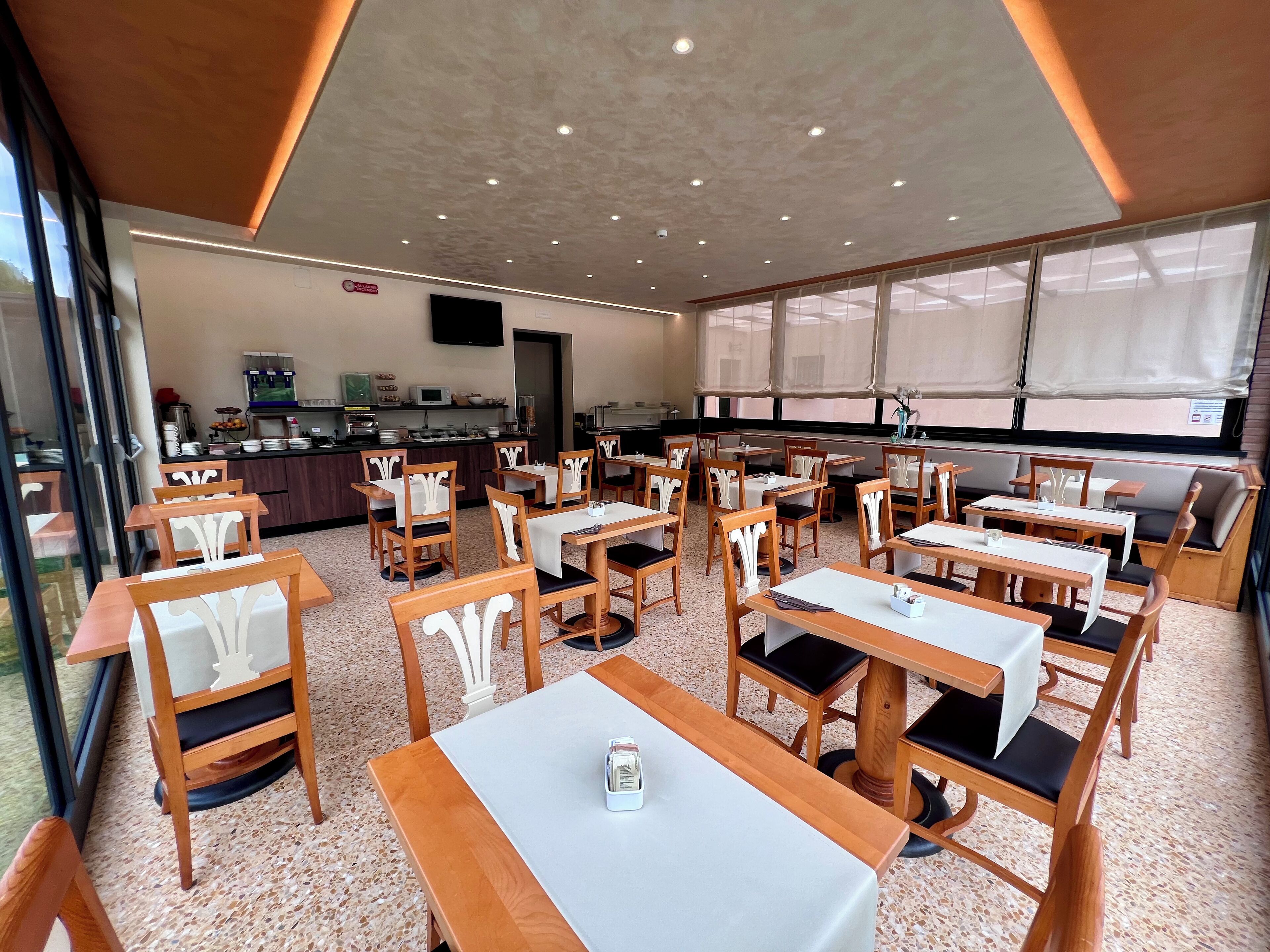 Un restaurant avec des tables et des chaises en bois, une cuisine à l’arrière-plan et de grandes fenêtres.
