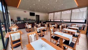 Un restaurant avec des tables et des chaises en bois, une cuisine à l’arrière-plan et de grandes fenêtres.